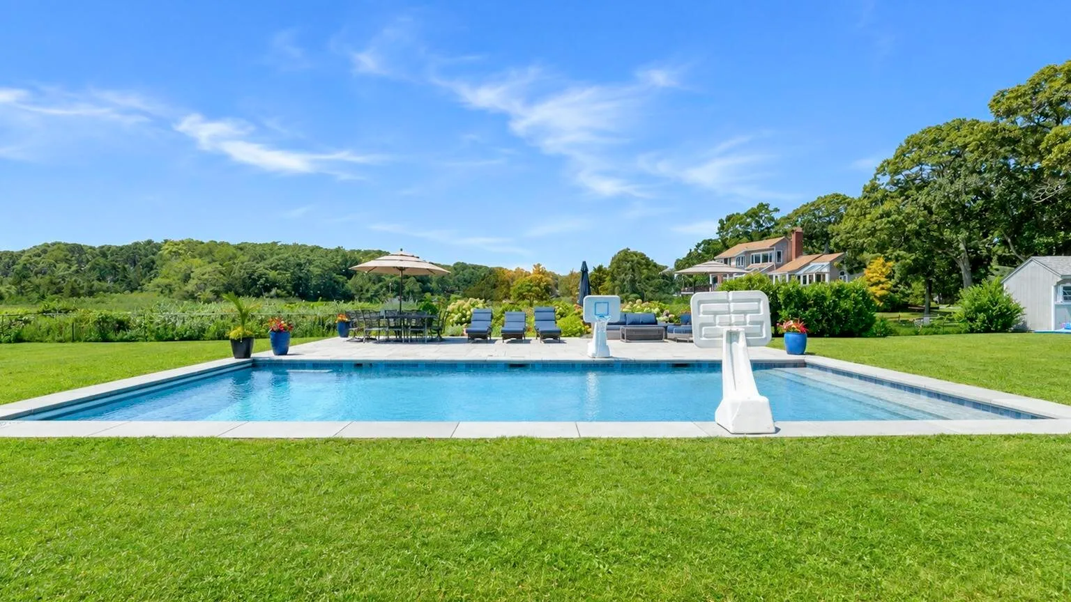 An outdoor swimming pool borders a broad green lawn, with a basketball hoop beside the water.
