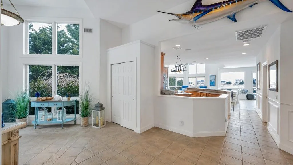 A bright open-plan interior has large windows, tiled flooring, and a kitchen beyond the counter.