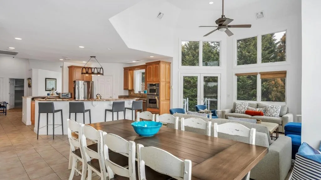 A bright open-plan dining and living area with large windows and a long wooden table.