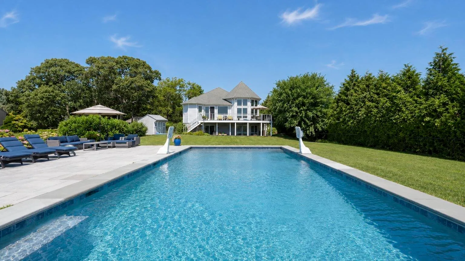 An outdoor swimming pool faces a large house, with lounge seating beside the water.