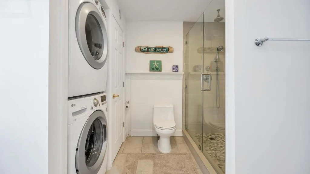 A bathroom with a toilet, glass shower, and stacked laundry machines.
