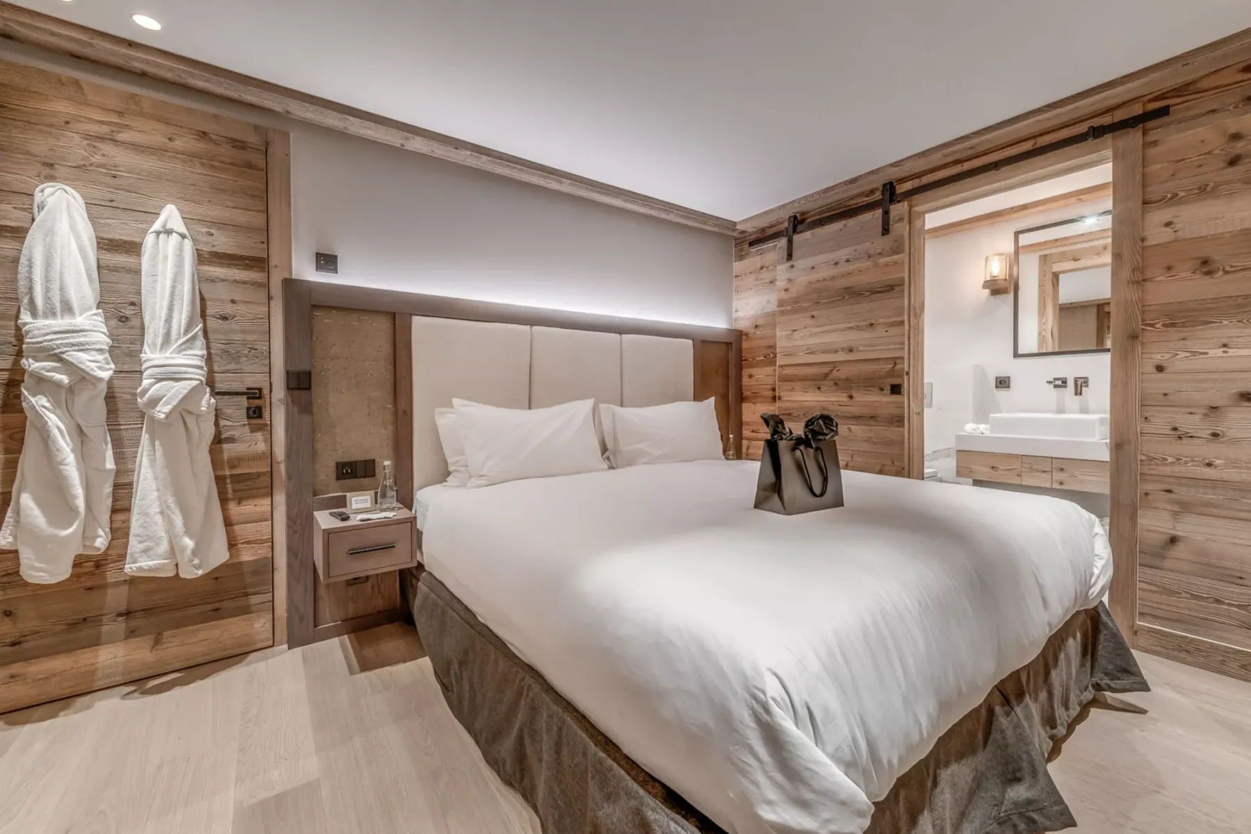 Un dormitorio con una cama tendida, paredes revestidas de madera y batas colgadas cerca.