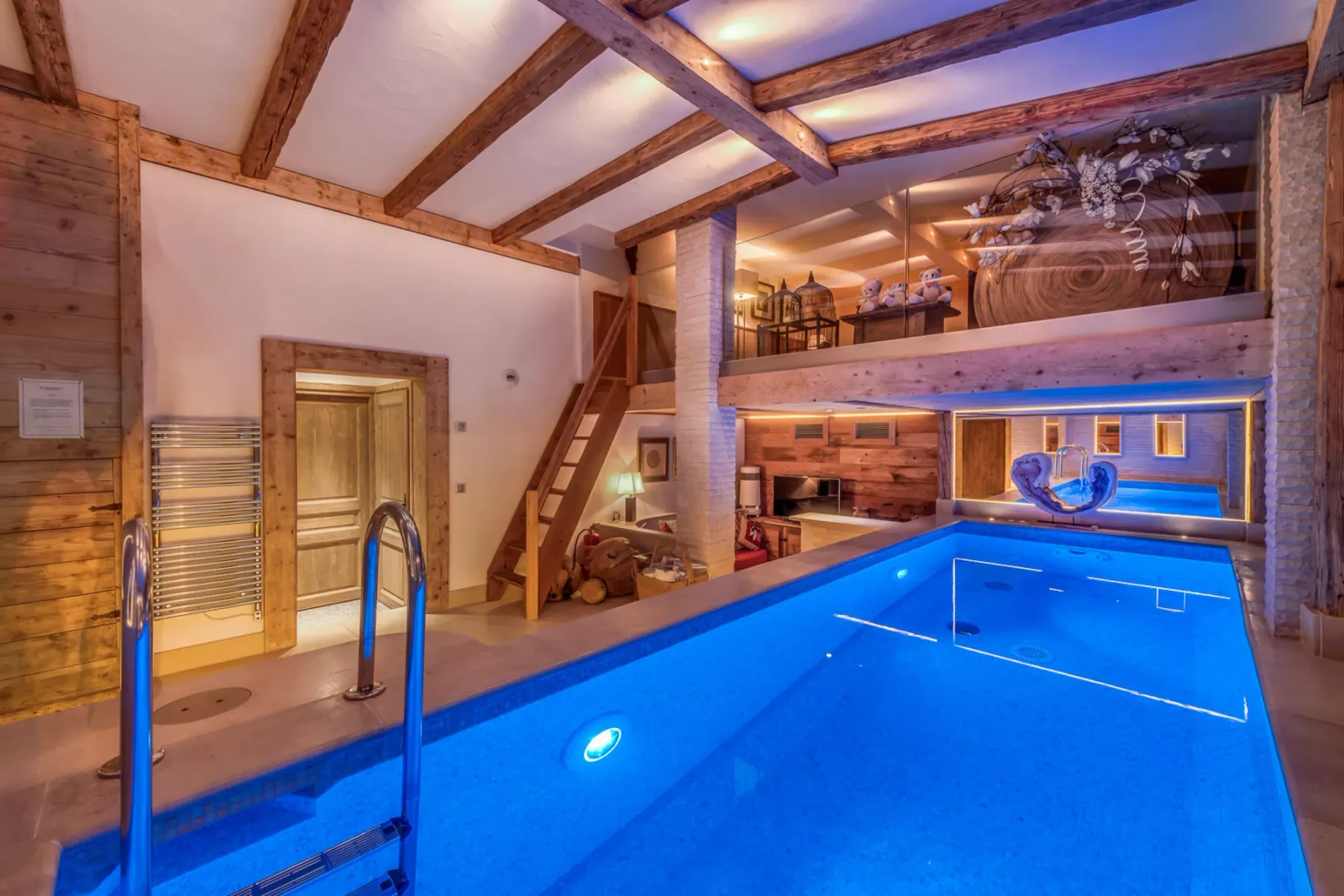 Una piscina cubierta está bajo vigas de madera expuestas, junto a una escalera y asientos en un nivel superior.
