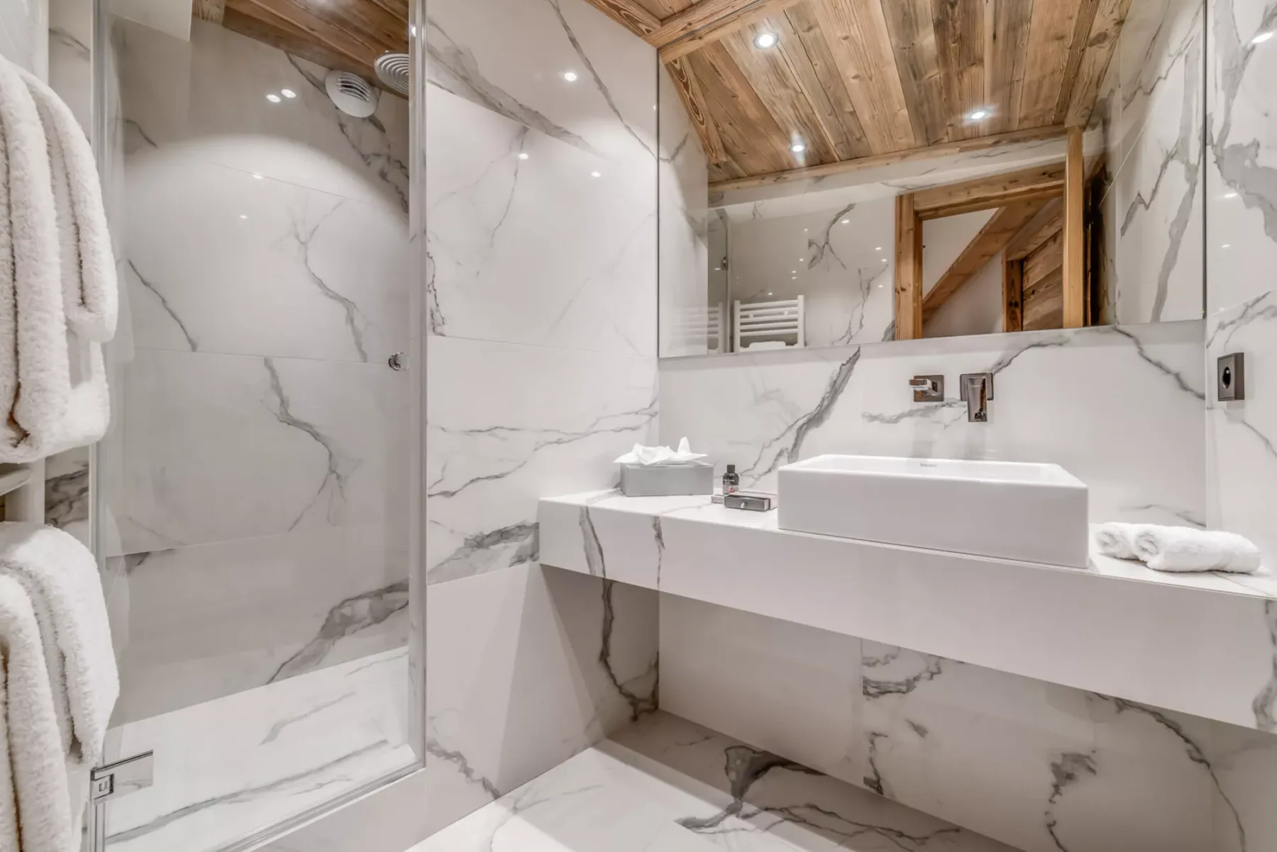Un baño revestido de mármol con ducha, lavabo sobre encimera y techo de madera.