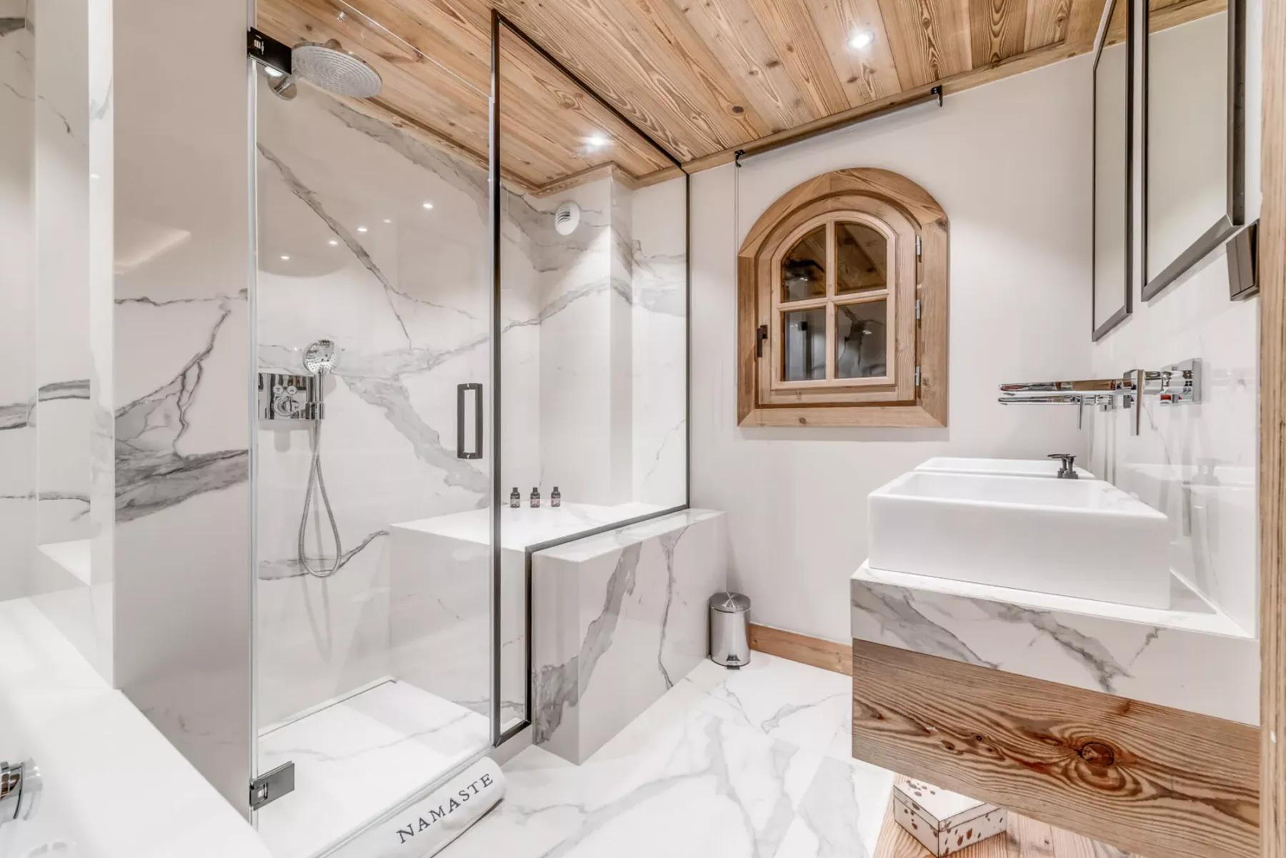 Un baño luminoso con ducha de vidrio, superficies con vetas de mármol y techo de madera.