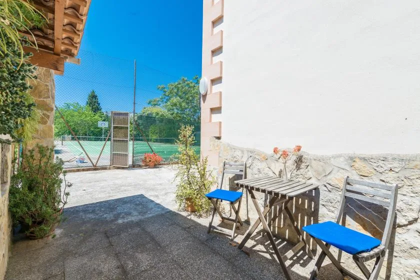 Un patio exterior con una mesa pequeña y asientos junto a una pista de tenis cercada.