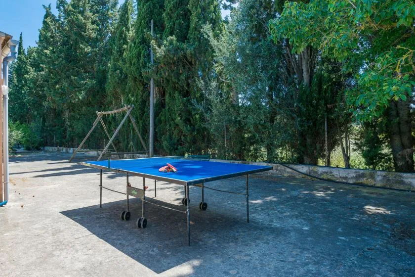 Una mesa de ping-pong al aire libre está en un patio pavimentado rodeado de árboles.