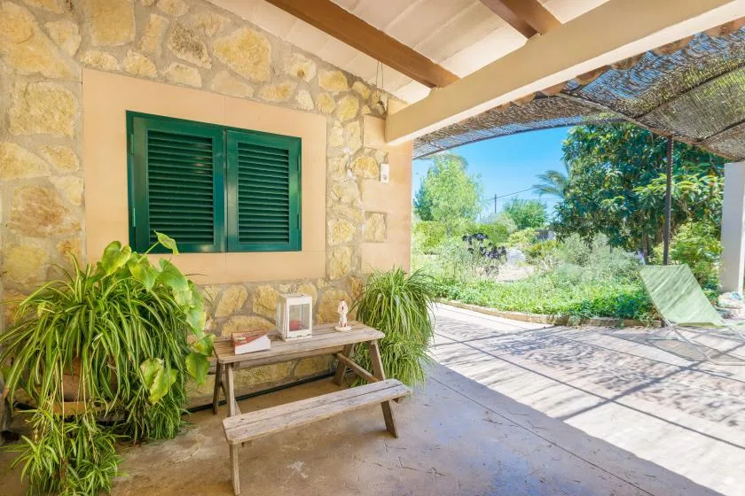 Un patio de piedra a la sombra, con un banco de madera y contraventanas verdes, da a un jardín.