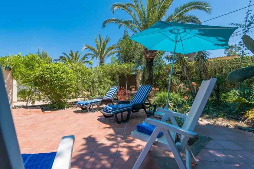 Un patio soleado al aire libre tiene asientos reclinables bajo una sombrilla turquesa.