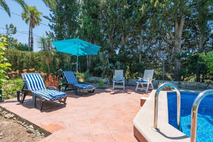 A poolside patio with lounge seating, a turquoise umbrella, and surrounding greenery.