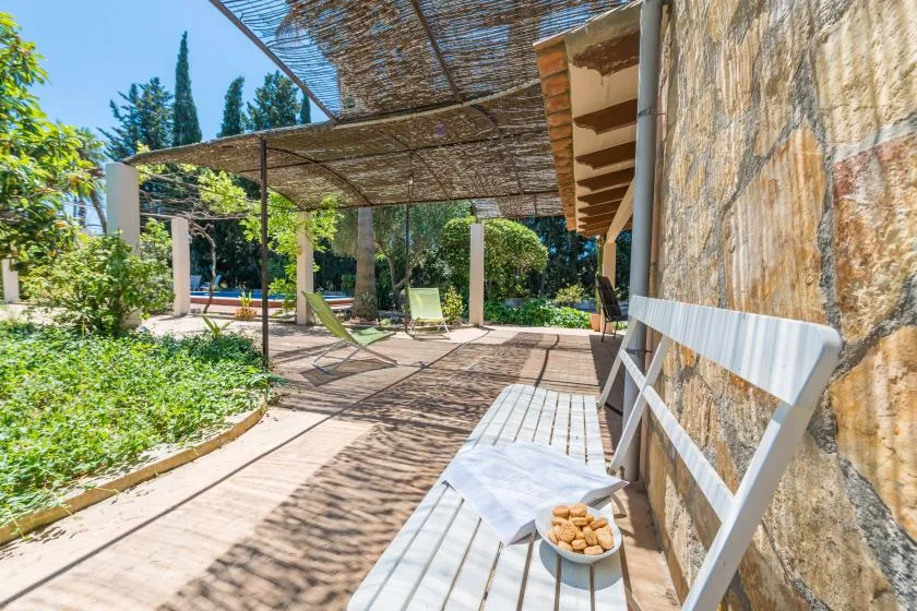 Un patio exterior con sombra, asientos y una mesa junto a un muro de piedra.