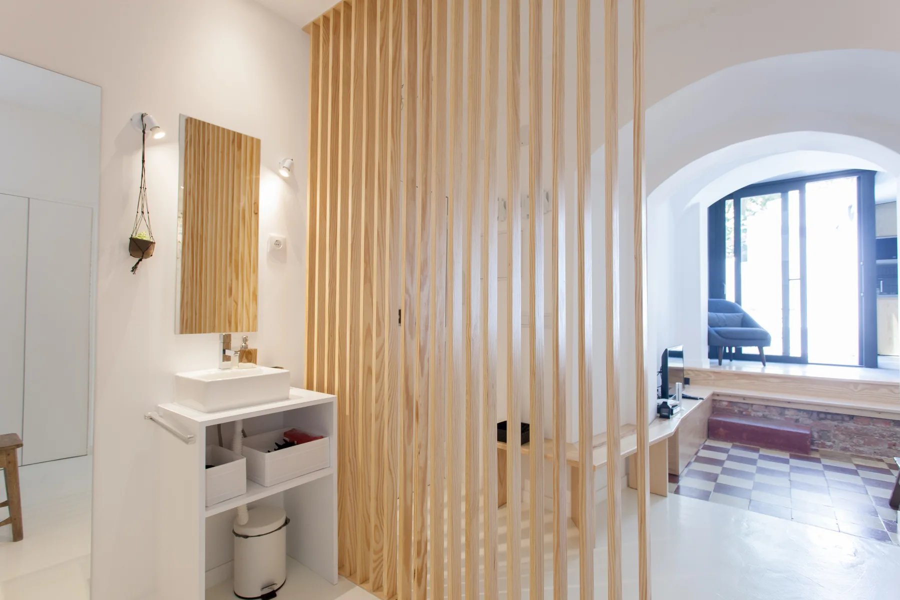 A bright bathroom with a sink and a wooden slat divider opening toward a sitting area.