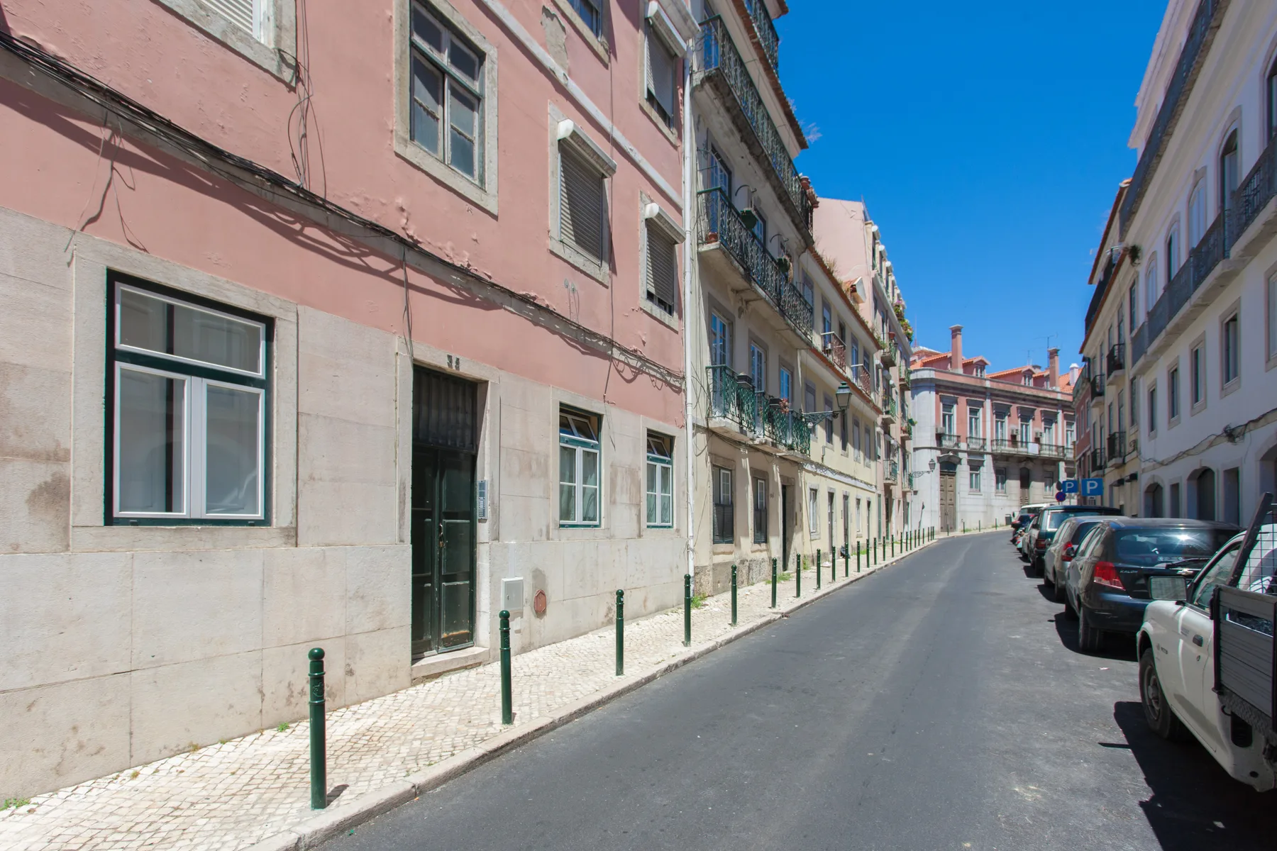 A narrow city street runs between tall buildings, with parked cars along the curb.