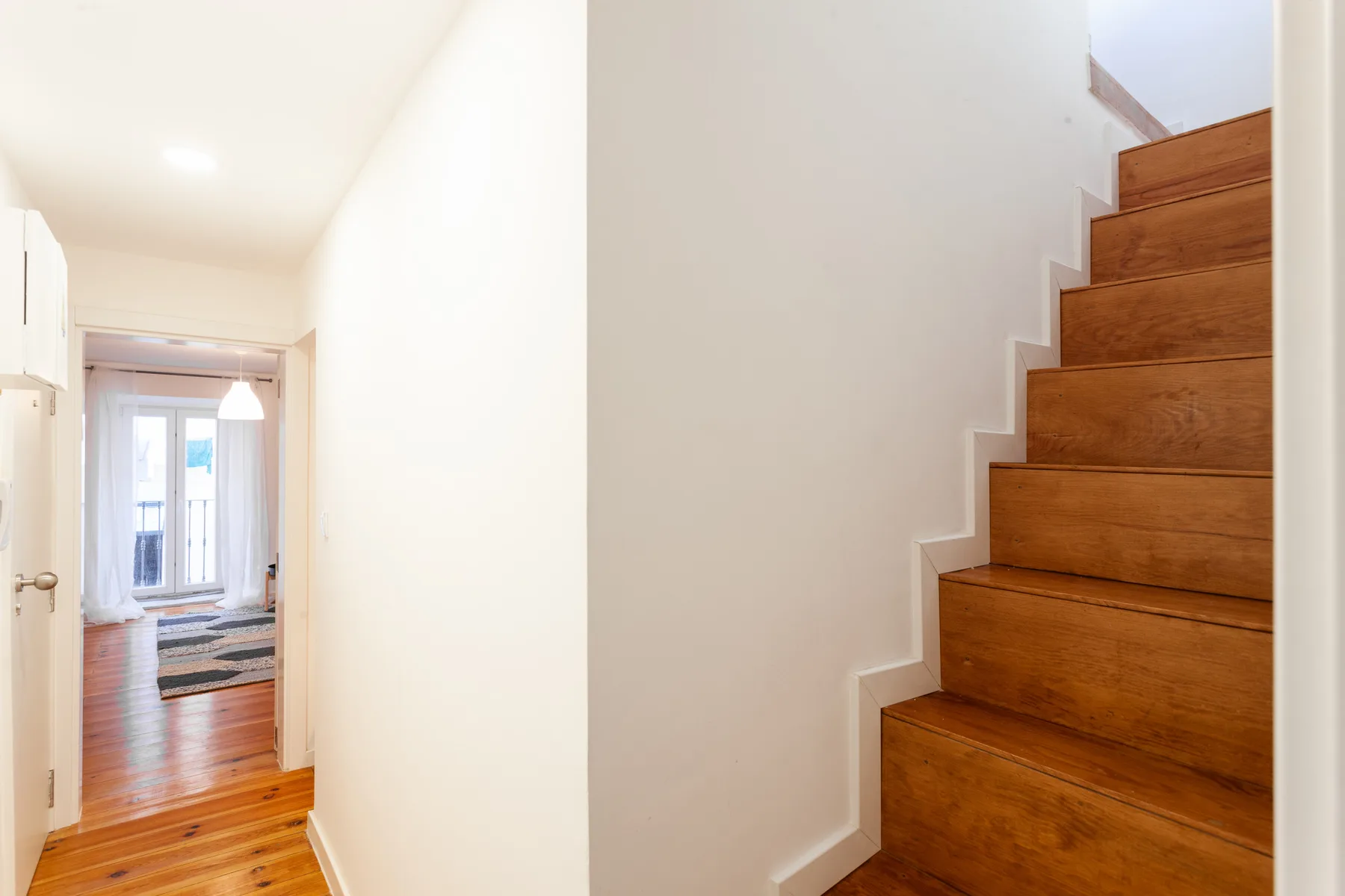 Un pasillo luminoso conduce a una escalera de madera y una puerta abierta.