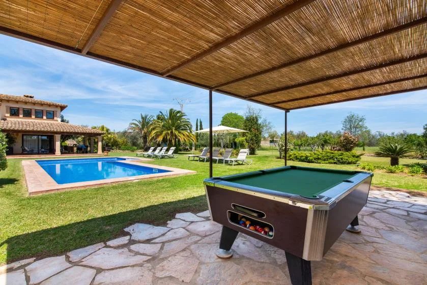 Zona recreativa al aire libre con piscina y futbolín bajo una cubierta de sombra.