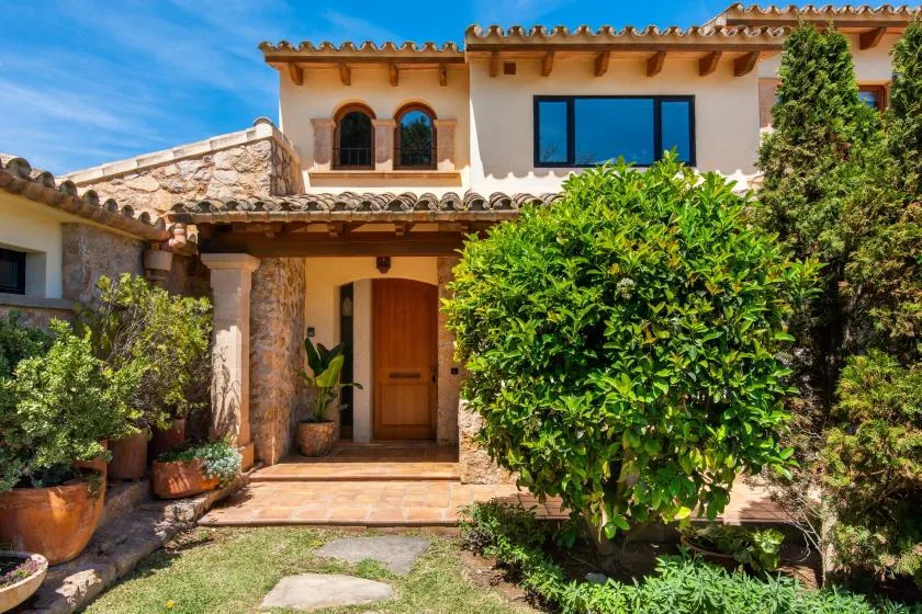 Exterior de una casa de estilo mediterráneo con puerta de madera y jardín frondoso.