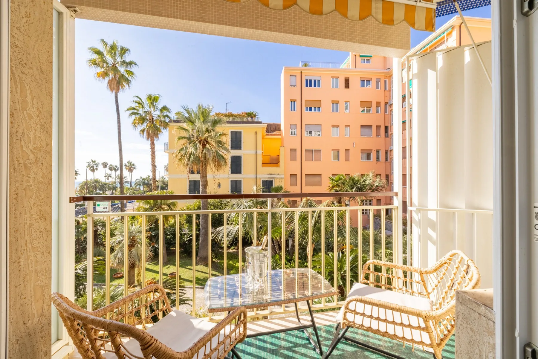A balcony with woven seating overlooks palm trees and nearby buildings.