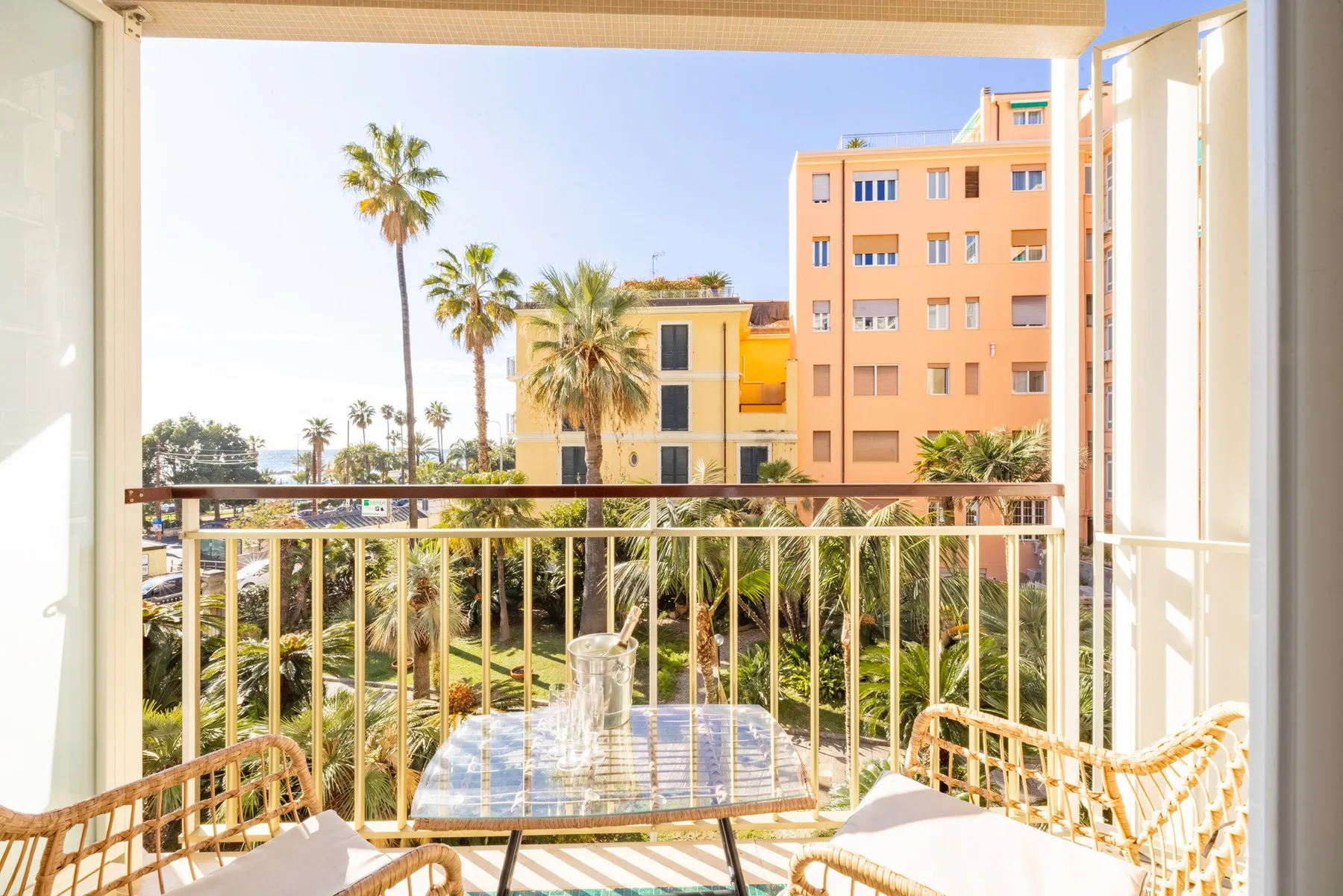 A balcony with seating overlooks palm trees and nearby apartment buildings.