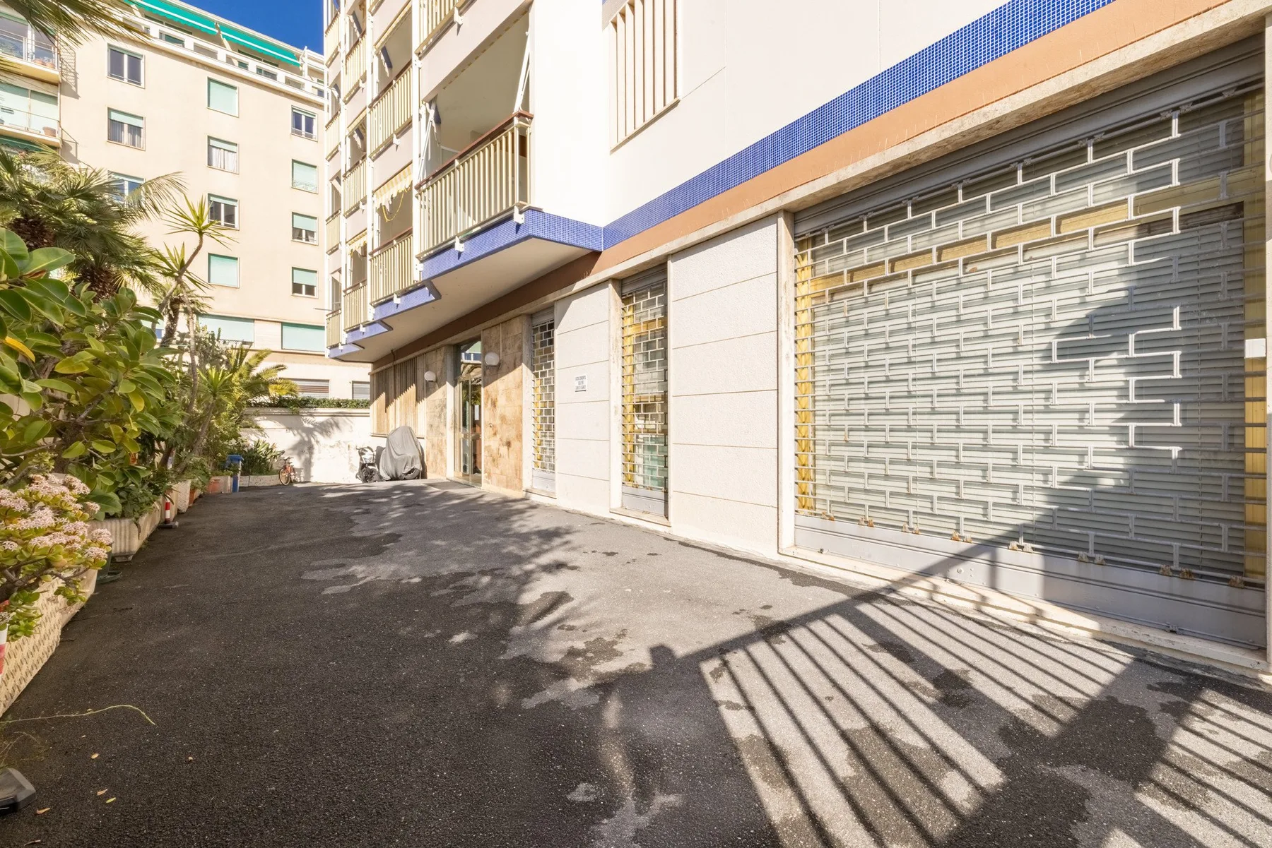 A paved courtyard runs beside a building with closed metal shutters and leafy plants.
