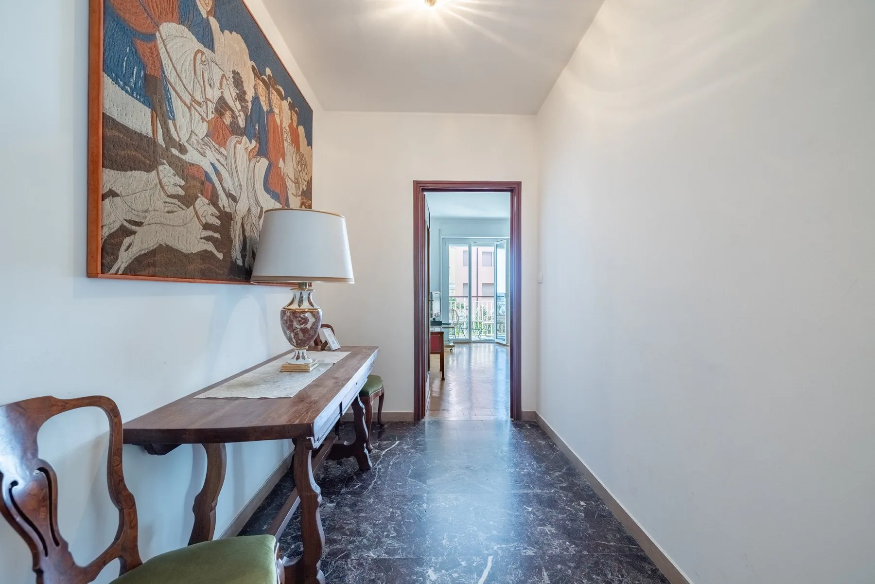A narrow hallway has a console table, a lamp, and a large wall painting.