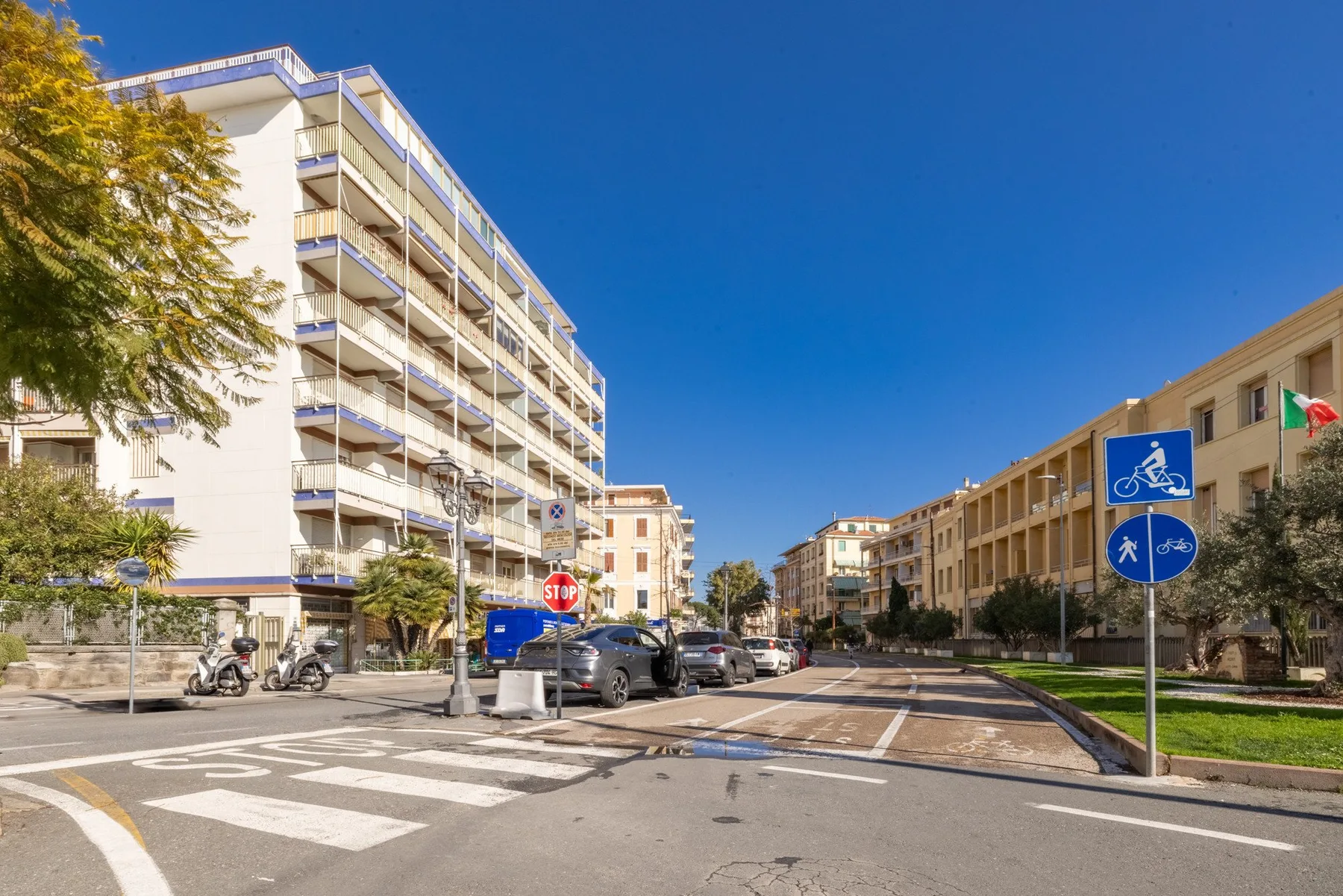 A sunny urban street with apartment buildings, parked cars, and a marked crosswalk.