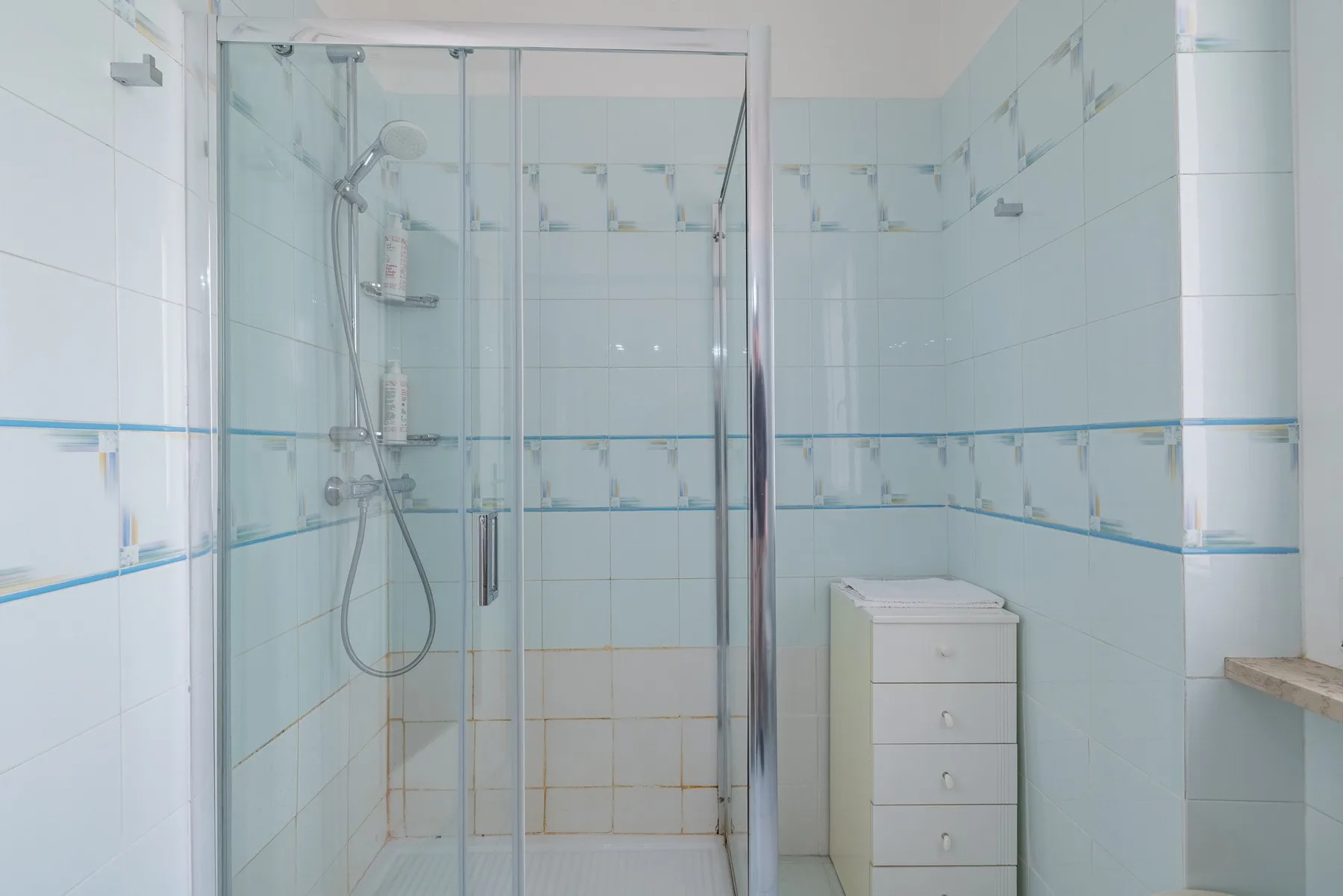 A tiled bathroom with a glass-enclosed shower and a small drawer unit.