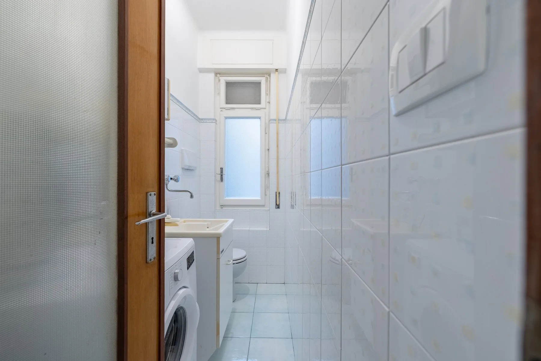 A narrow tiled bathroom with a sink, washing machine, and frosted window.