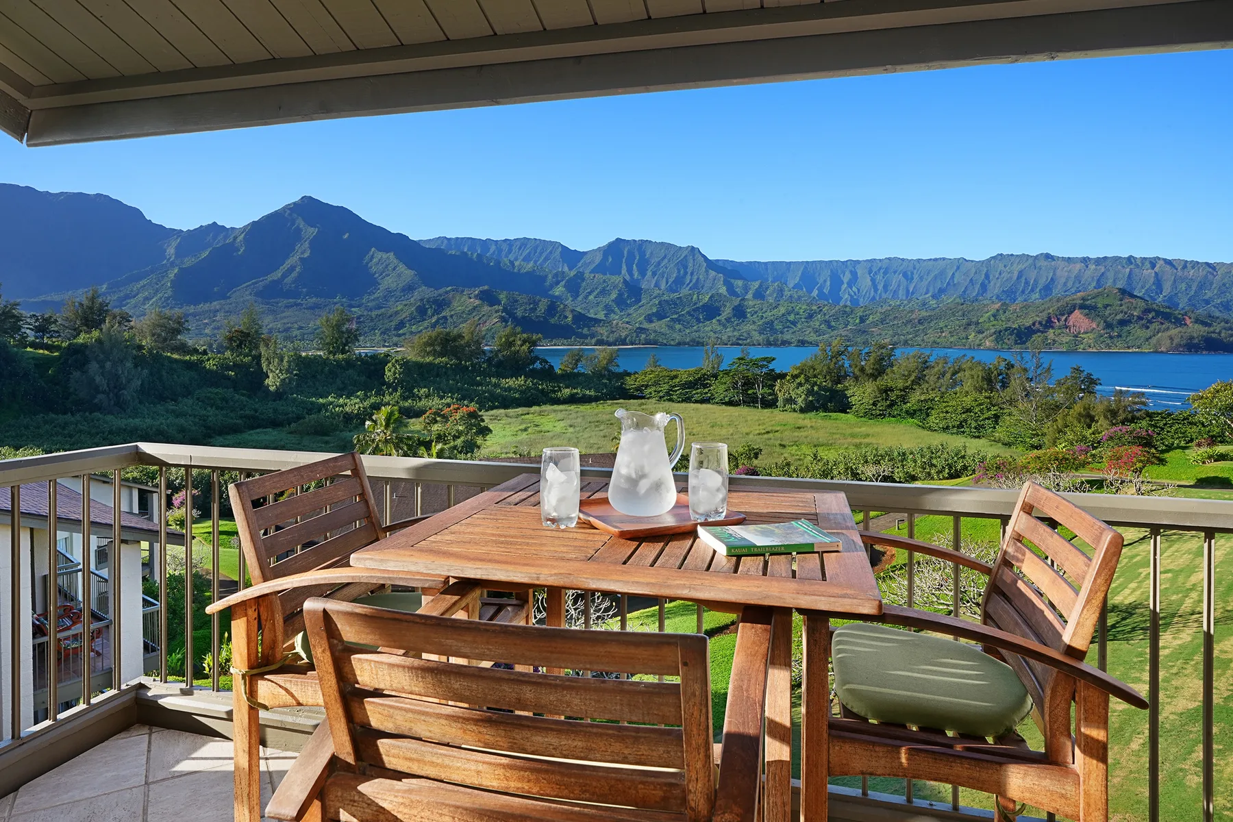 A covered outdoor patio has wooden seating around a table overlooking a lake and mountains.