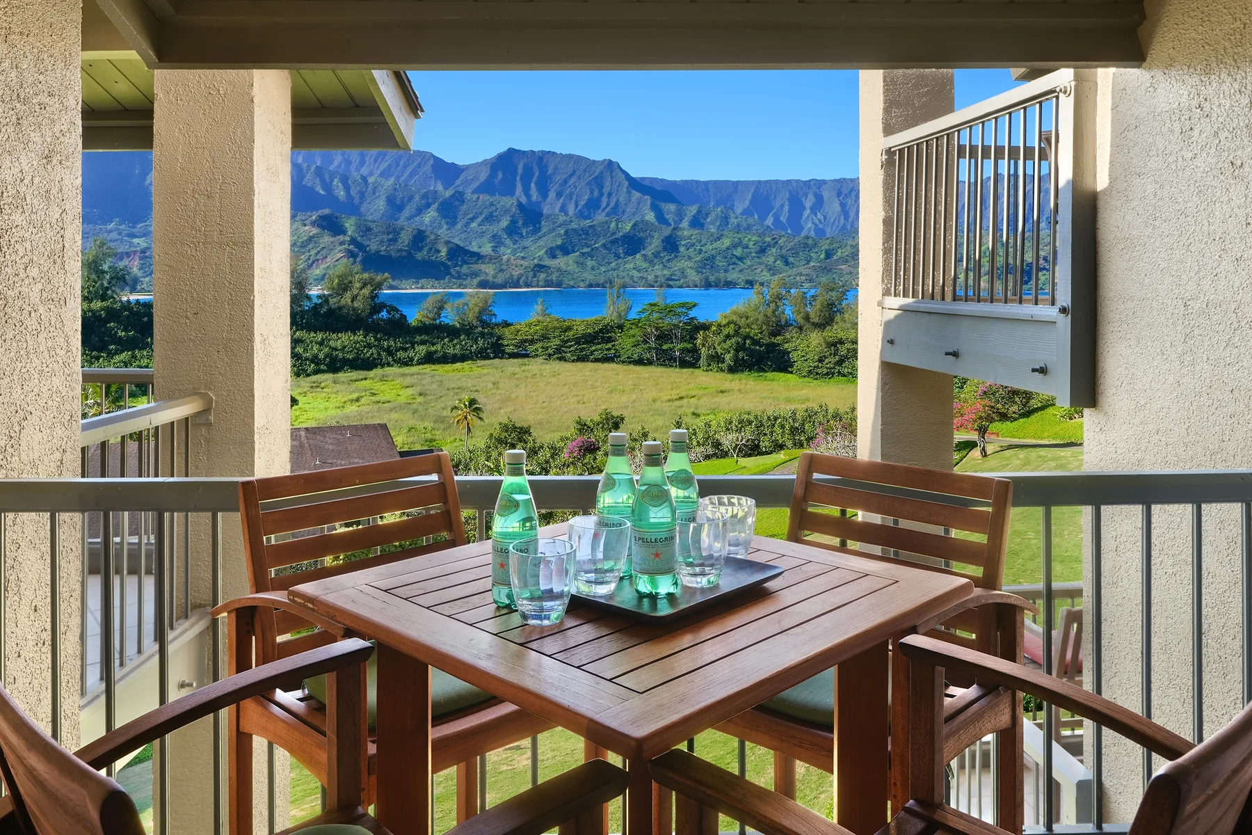 A covered balcony dining area overlooks a green landscape, blue water, and mountains.