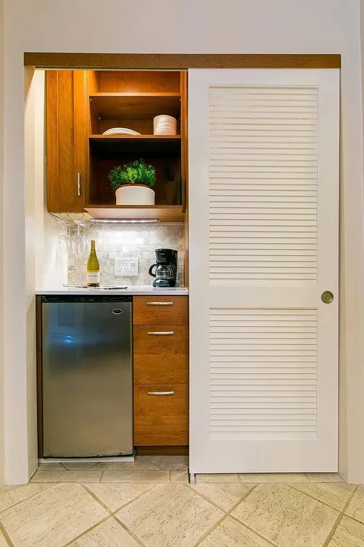 A compact kitchenette with wooden cabinets, a small refrigerator, and a louvered door.