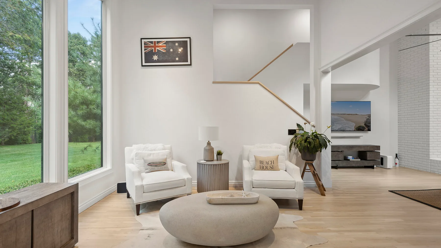 Una sala luminosa con dos sillones y un otomano redondo junto a grandes ventanas.