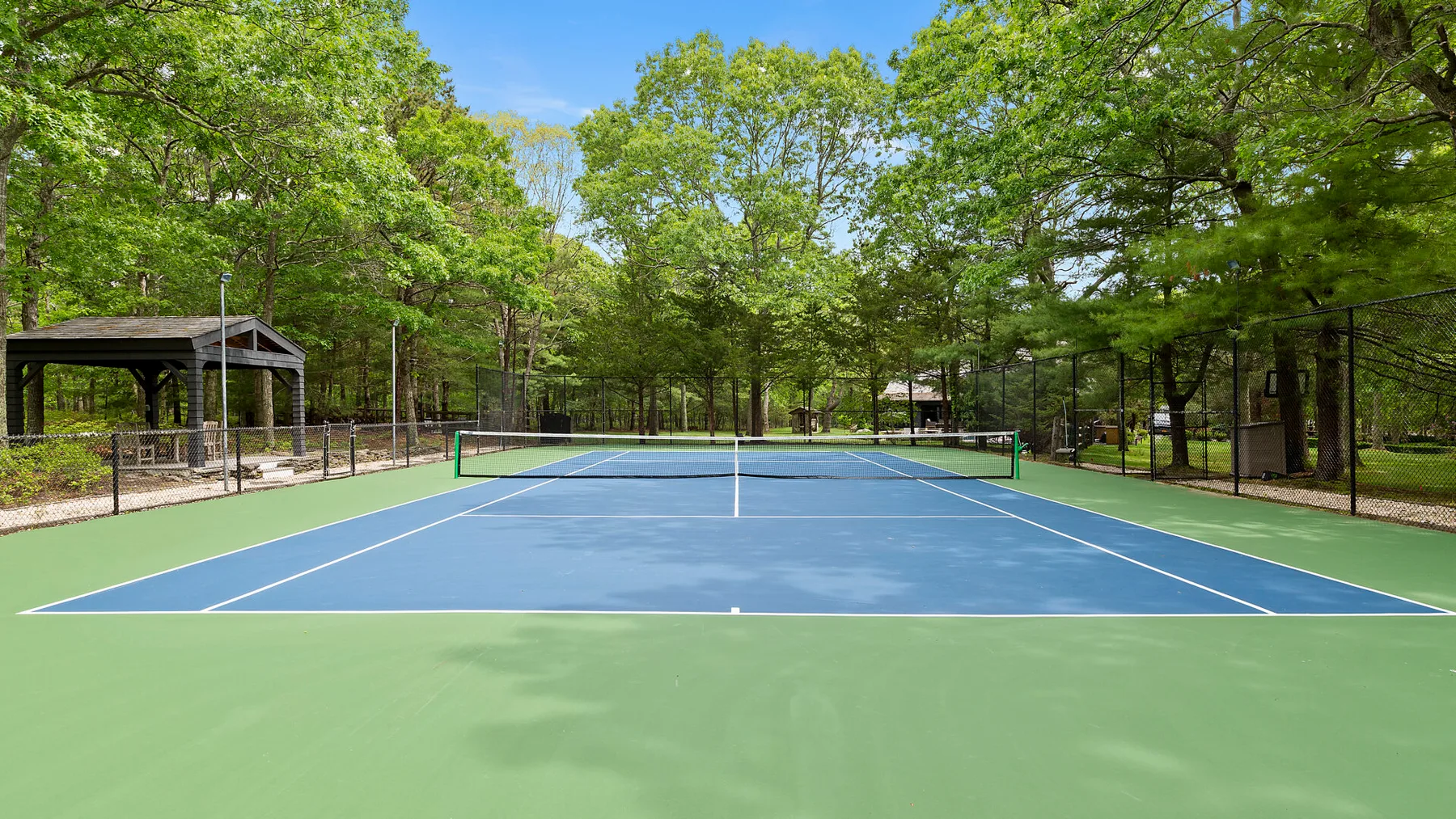 Una cancha de tenis al aire libre está rodeada de árboles frondosos y tiene un refugio sombreado al lado.