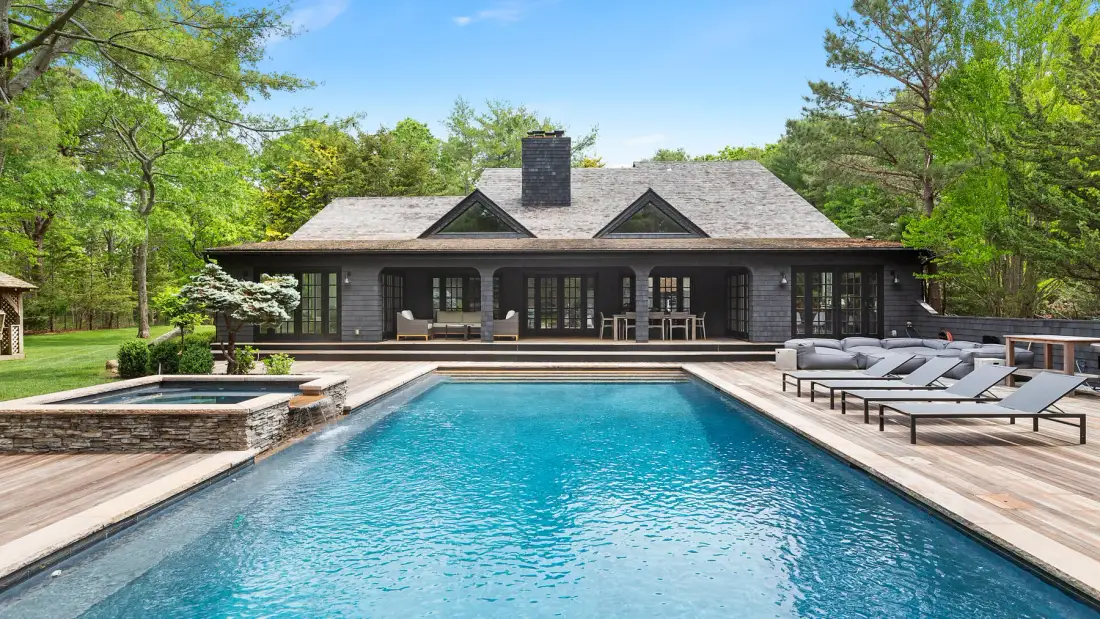Una piscina exterior está frente a una casa oscura, con asientos junto a la terraza.