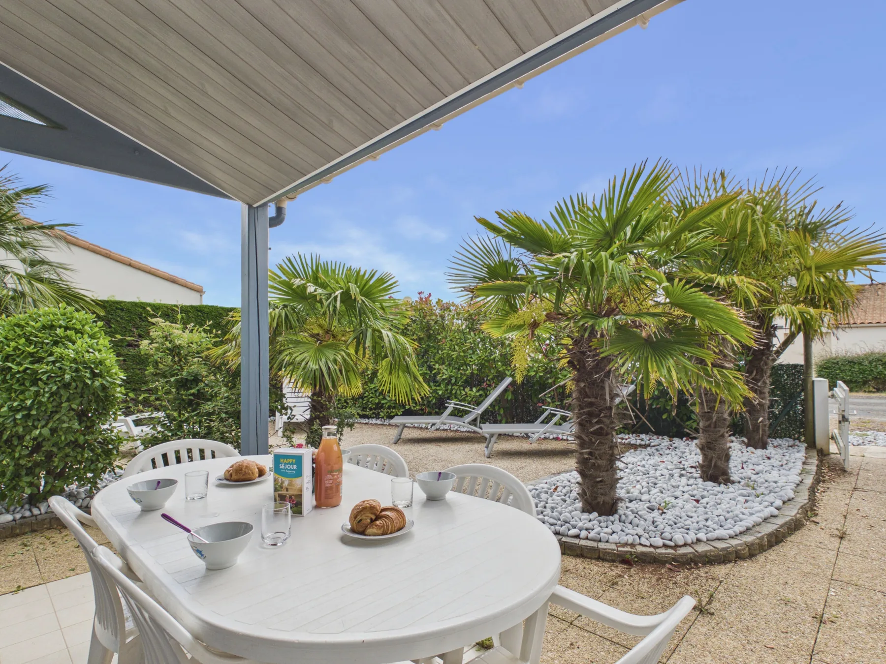 A shaded outdoor dining area overlooks a garden with palm trees.