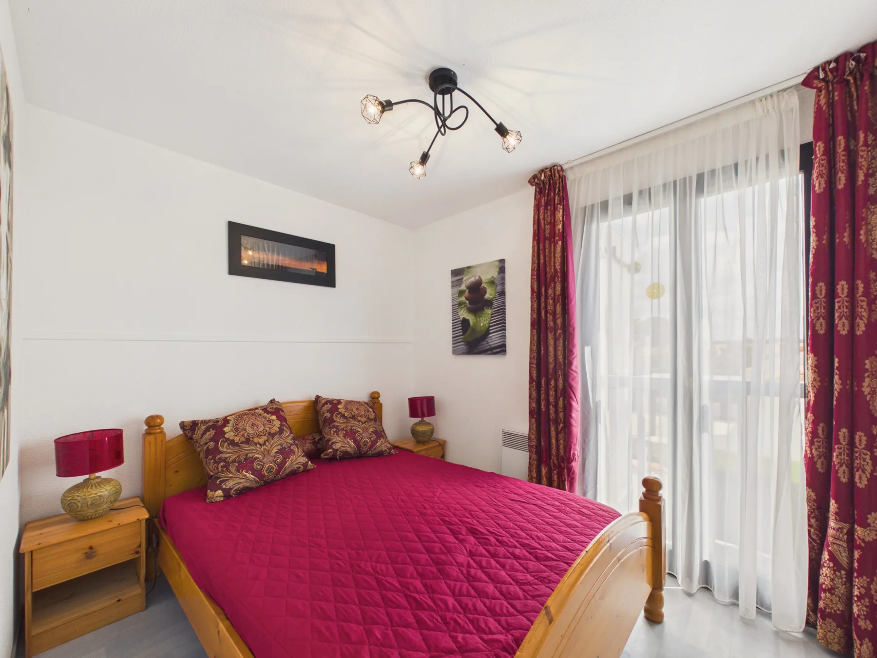 A bedroom with a bed covered in a deep red quilt beside curtained windows.