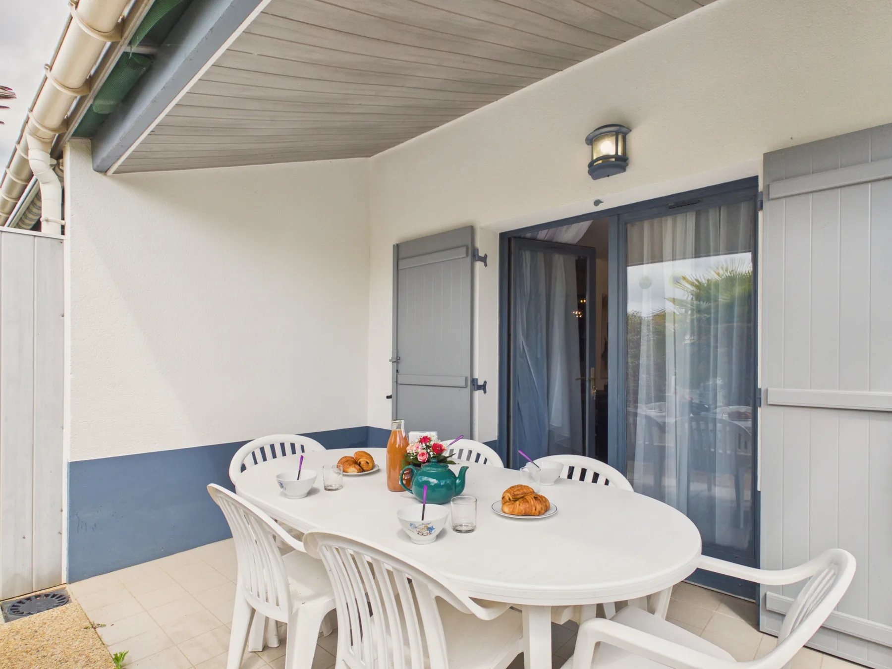 A covered patio with an outdoor table and white seating beside glass doors.