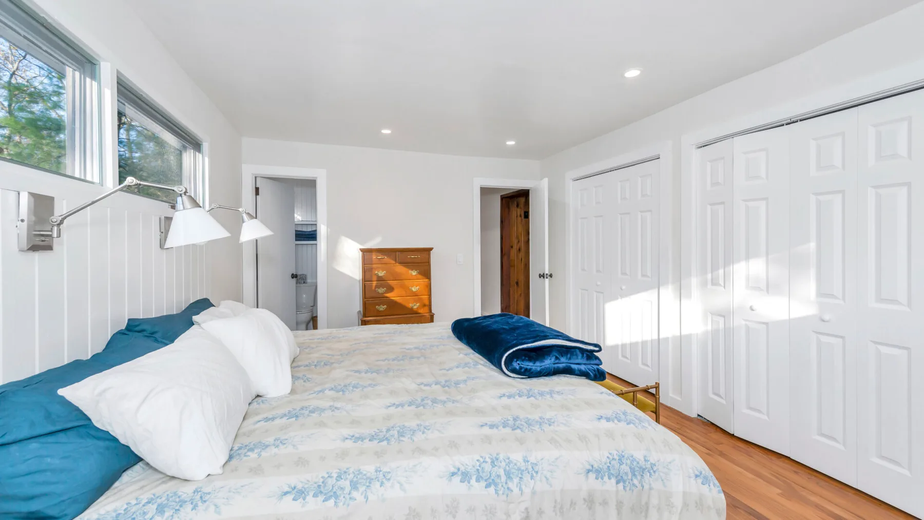 Un dormitorio luminoso con una cama, puertas blancas con paneles y ventanas en una pared.