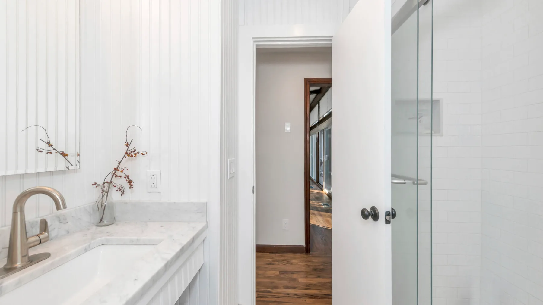 Un baño luminoso con un lavabo de mármol y una ducha de vidrio junto a una puerta abierta.