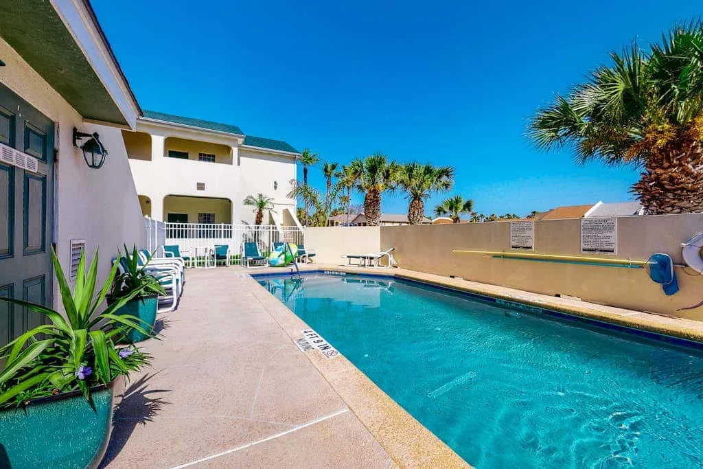 Una piscina al aire libre junto a una terraza pavimentada, con asientos y palmeras.