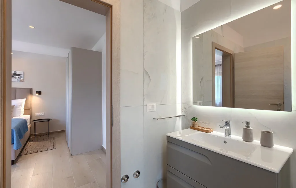 Un tocador de baño luminoso con un espejo amplio, junto a una puerta abierta a un dormitorio.