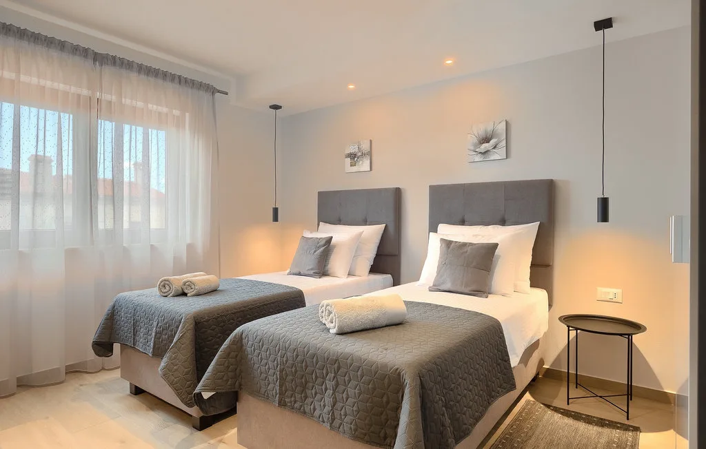 Un dormitorio luminoso con dos camas, ropa de cama gris y cortinas translúcidas.