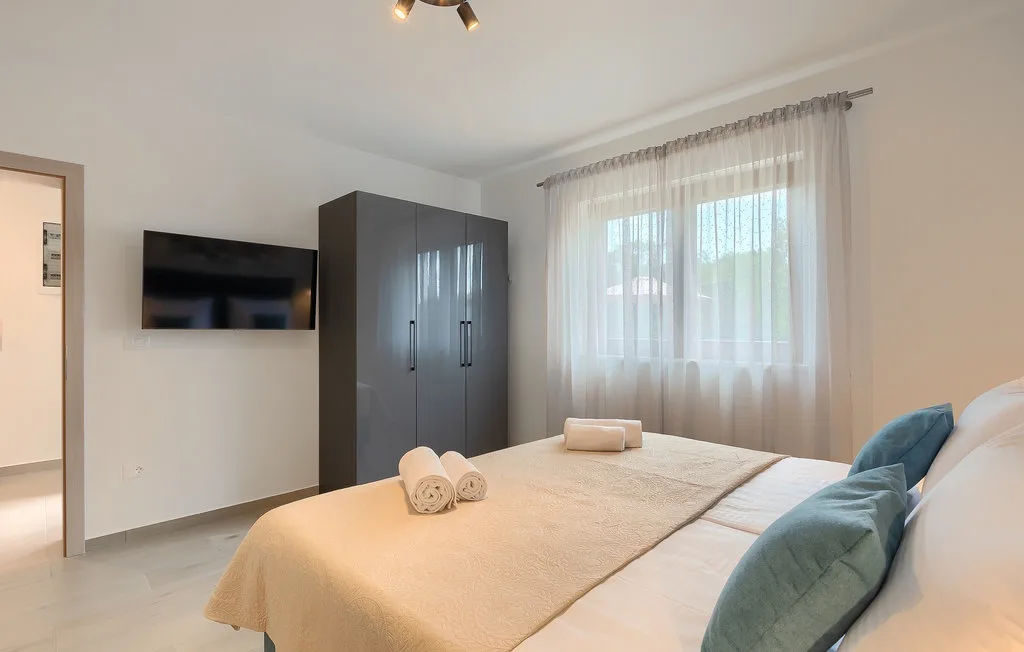 Un dormitorio con una cama bien hecha, un armario oscuro y una ventana con cortinas translúcidas.