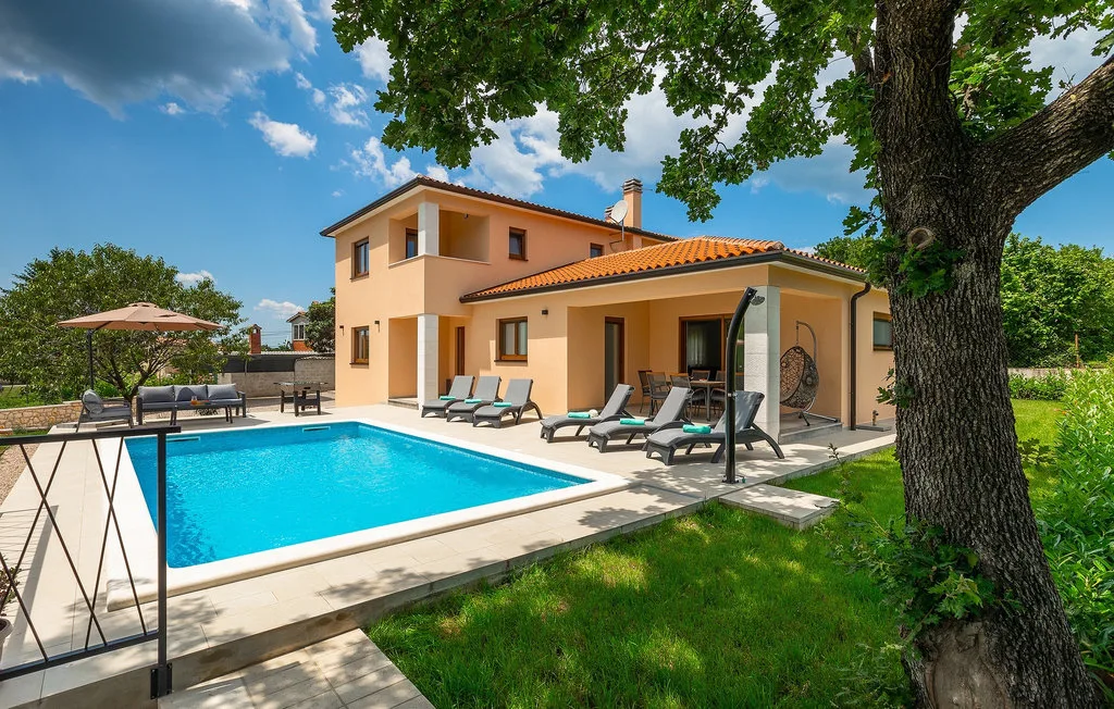 Una casa y una piscina exterior se encuentran bajo árboles frondosos en un jardín soleado.