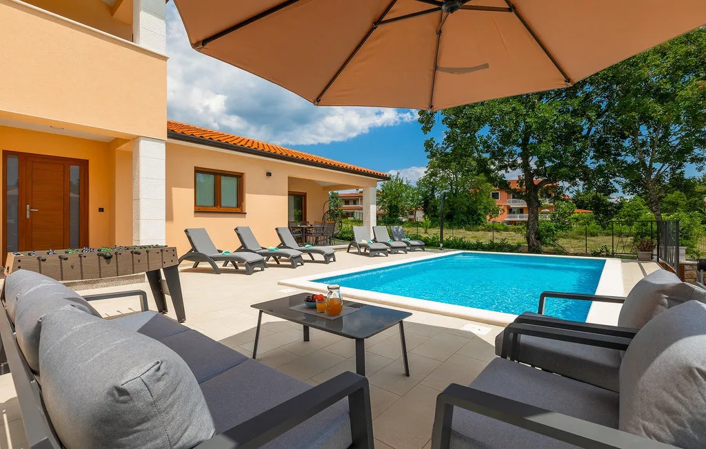 Una piscina junto a un patio con asientos reclinables y una sombrilla grande.