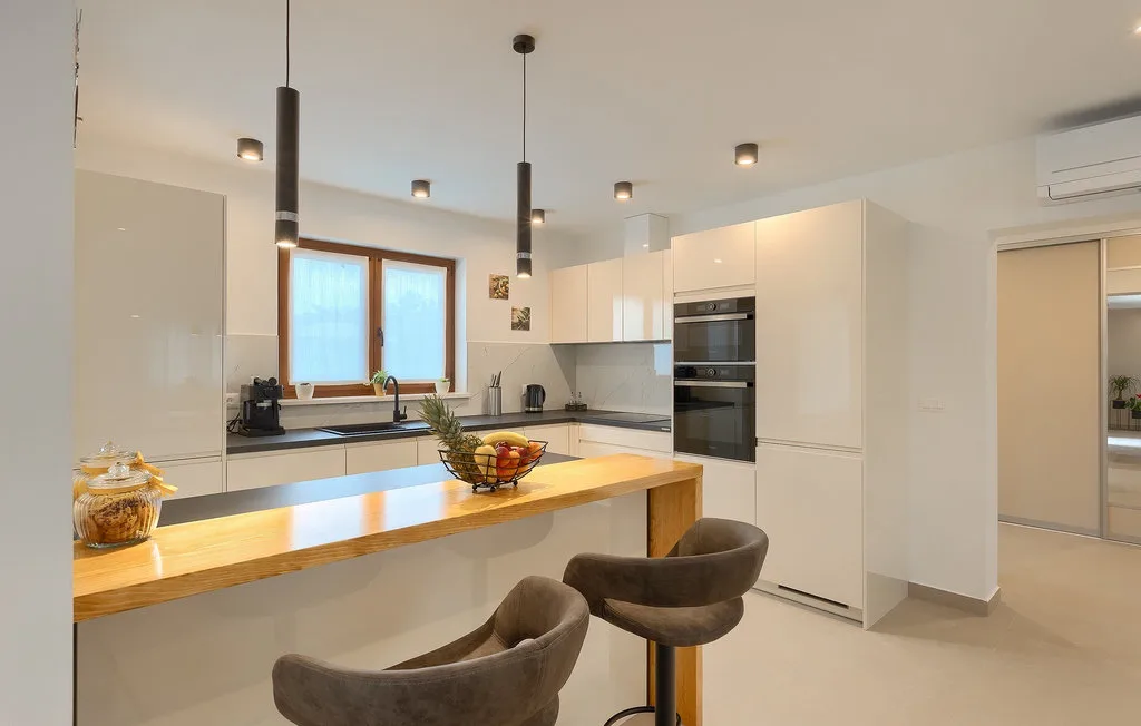 Una cocina luminosa con gabinetes blancos, una barra de madera y lámparas colgantes.