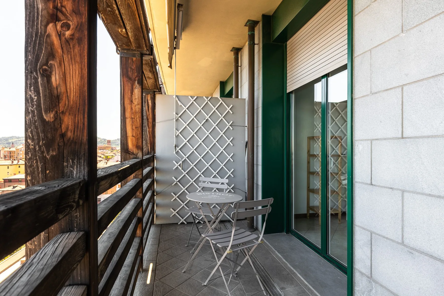 Un balcón estrecho con una mesa pequeña y sillas junto a puertas de vidrio.