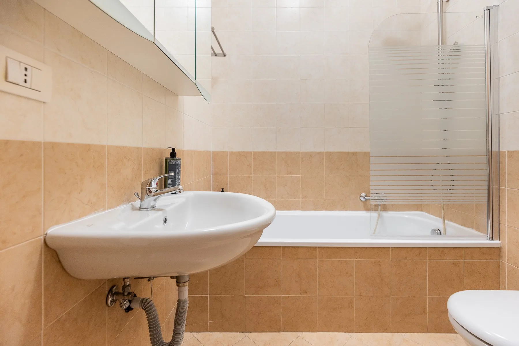Un baño con azulejos, lavabo de pared y bañera con una mampara de vidrio.