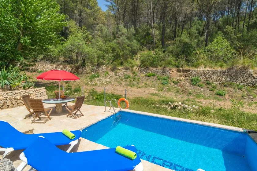 An outdoor swimming pool beside a patio with seating and a red umbrella.
