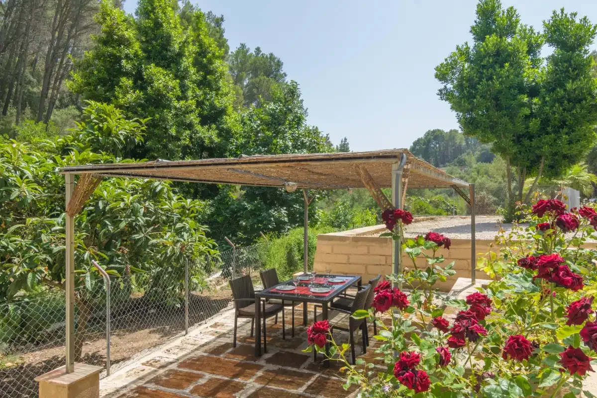 Una zona de comedor al aire libre bajo una pérgola de madera, rodeada de vegetación y flores.