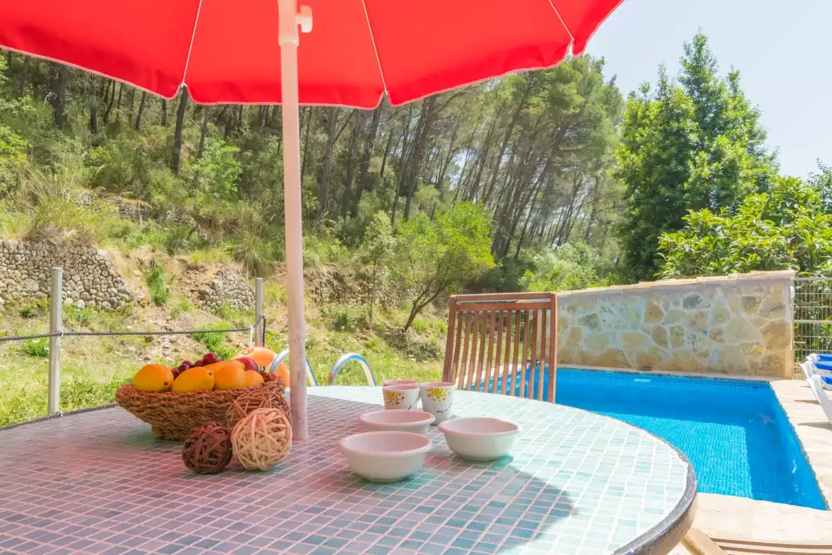 Una mesa exterior de azulejos con fruta está junto a una piscina bajo una sombrilla roja.